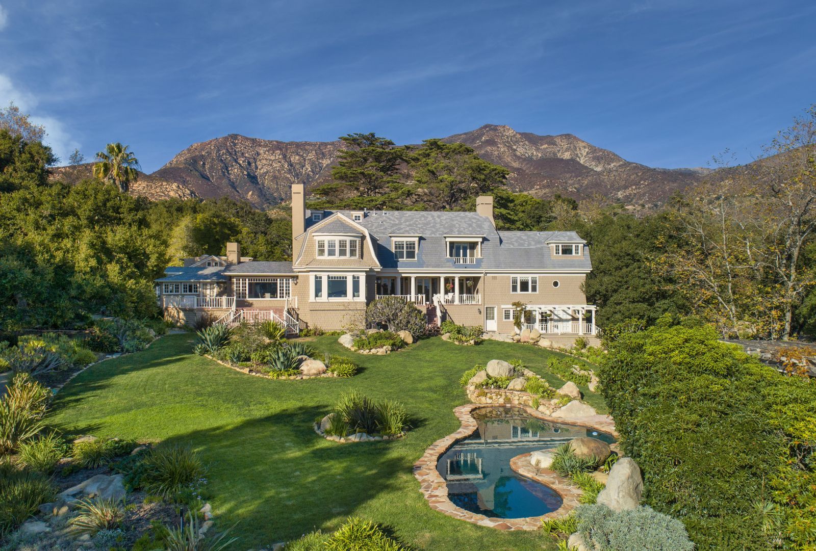 A Cape Cod-style home and its lush grounds with mountains in the background and a serene blue pool in the foreground.